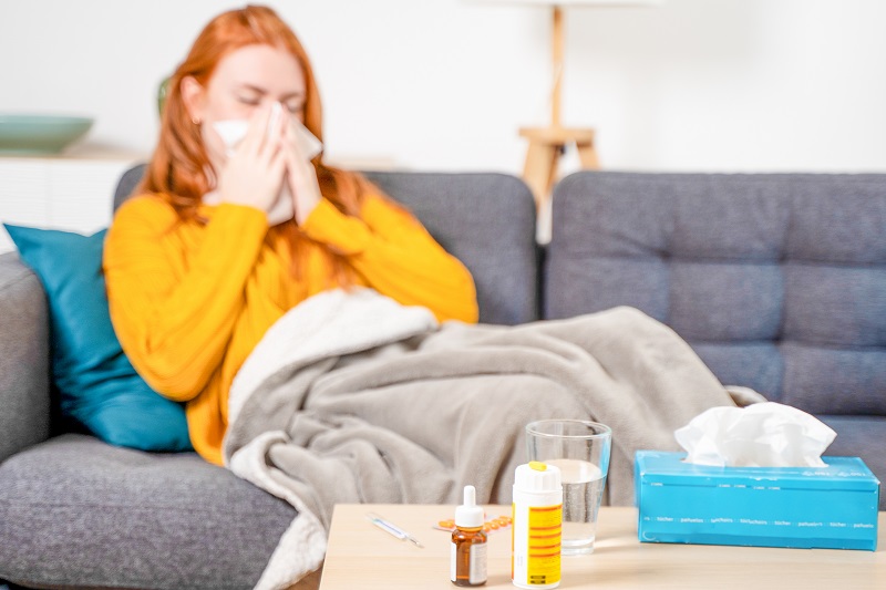 Woman home sick with the flu, blowing her nose on the couch. Over-the-counter pain relievers and medicine on the coffee table.