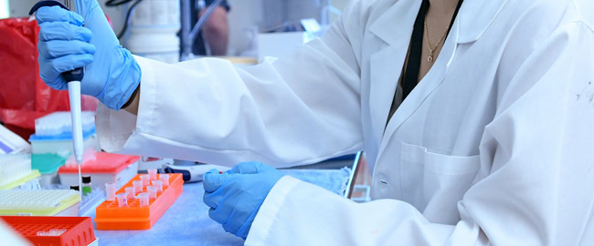 Person in white lab coat and blue rubber gloves using an eyedropper in medicine tube