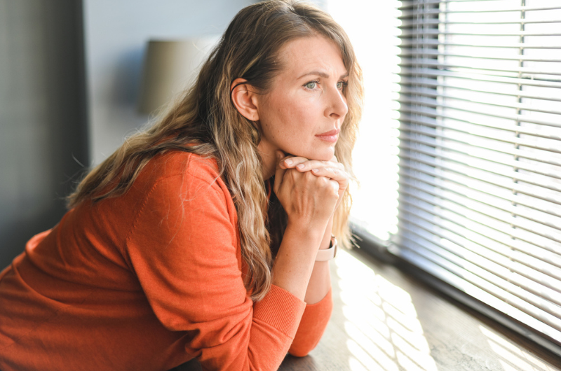 A mature adult woman rests her chin on her hands and looks out the window.