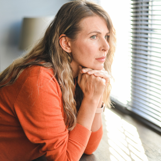 A mature adult woman rests her chin on her hands and looks out the window.