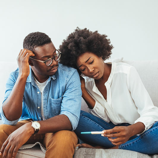 A sad young woman holds a pregnancy test in her hand, sitting on the couch with her partner.