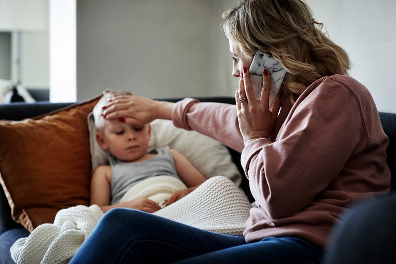 Mother touches her son's forehead as he rests on the couch, feeling for a fever. Young boy not feeling well, possibly sick with RSV.