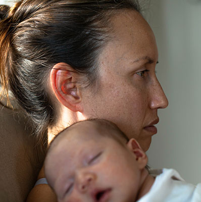 A mom experiencing the "baby blues", feeling unhappy, holding her baby at home sitting on the couch thinking.