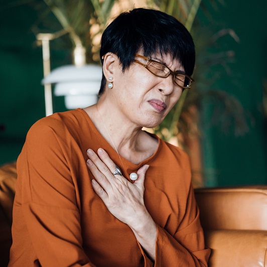 Woman with eyes closed holding her chest in discomfort, suffering from chest pain while sitting on the sofa at home.