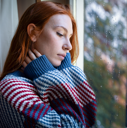 Woman looking out the window depressed as it rains and is darker outside, experiencing seasonal affective disorder.