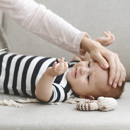 A mother's hand touching her babies forward to feel if it it is warm, while holding a thermometer reading the baby's temperature with a fever of 100 degrees