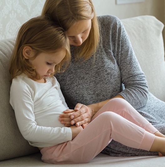 Child sitting on sofa and suffering from constipation, mother touching her belly trying to offer comfort.