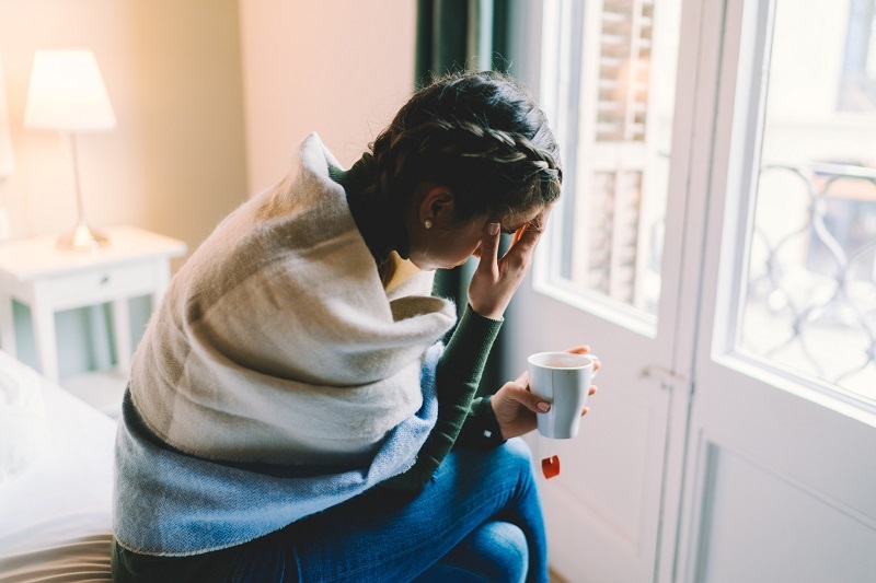 Young woman, sitting on the edge of the bed, bent over with her hand on her face, struggling with depressive symptoms from premenstrual dysphoric disorder.