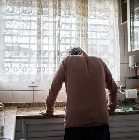 From behind, an older man leaning on a counter with fatigue, not feeling well.