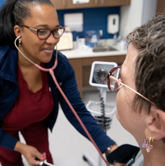 Woman at a primary care appointment, with a nurse leaning in to listen to her heart with a stethoscope.