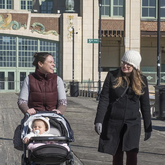 Three friends walking on the Asbury Park boardwalk, smiling, and pushing strollers with their children.