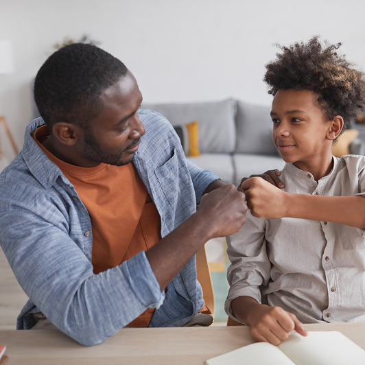 A father and school-aged son fist-bumping and smiling in their home.