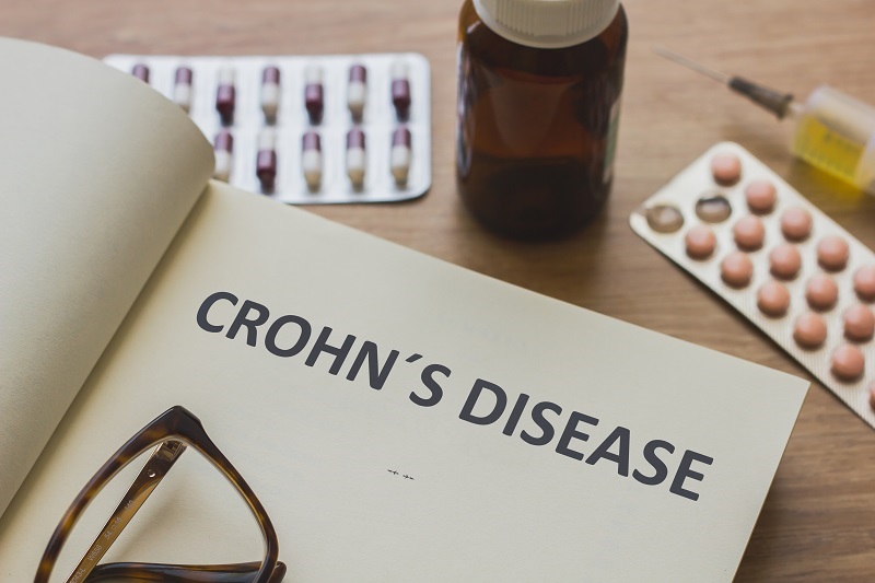 A book opened that says "Crohn's Disease" on the cover page. Around the book are reading glasses and some medications.