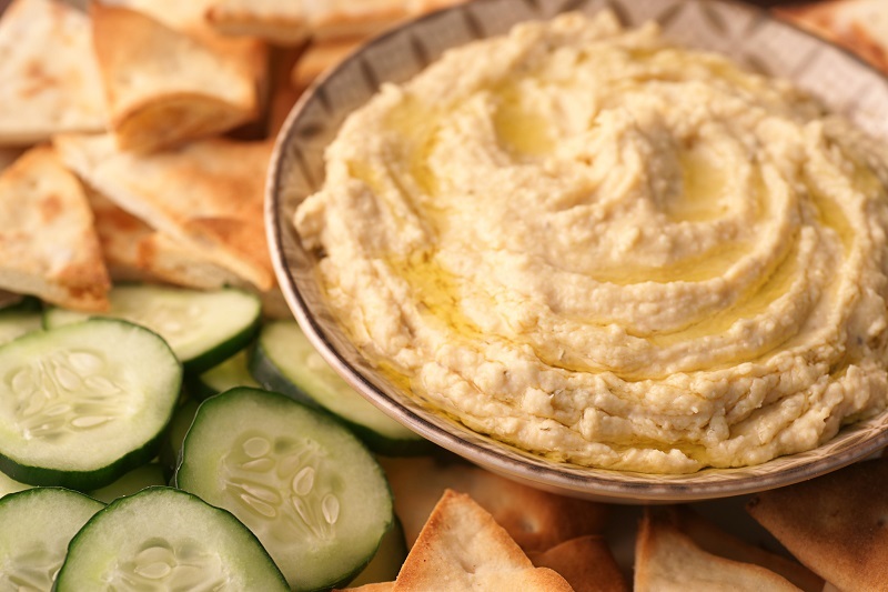Lemon, garlic and white bean hummus in a bowl with sliced cucumbers and pita surrounding it.