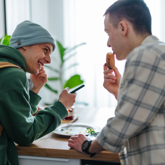 Two teenage boys eat food off a plate while smiling and laughing.