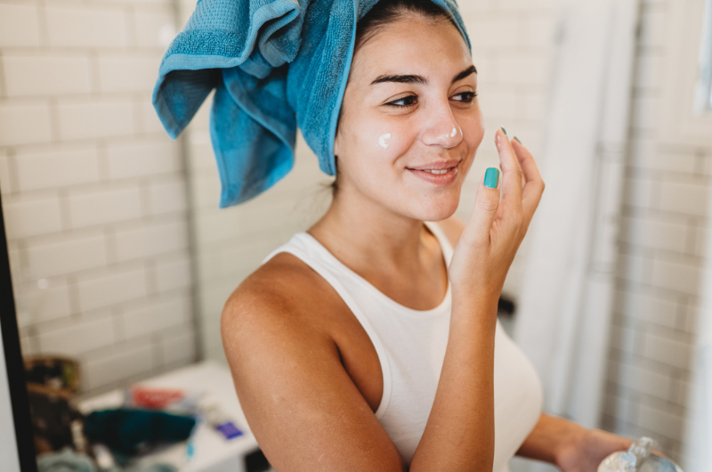 A young woman with her hair in a towel dabs a skincare product on her face.
