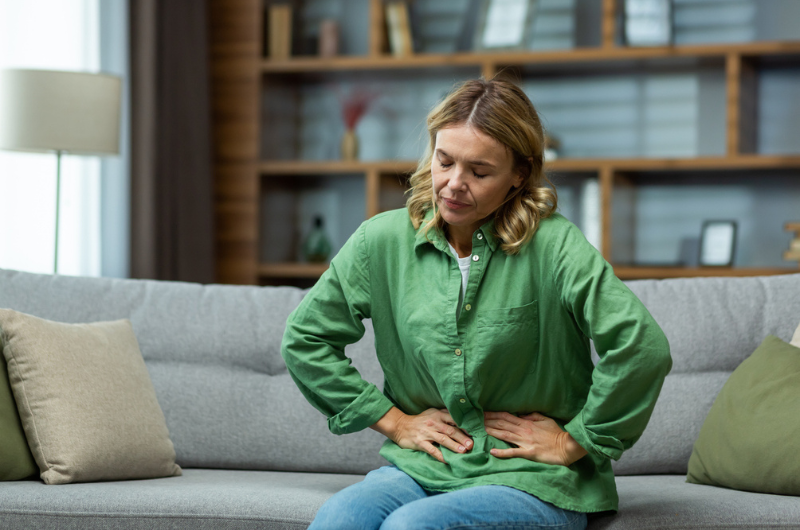 A woman is sitting on the sofa at home, holding her stomach with her hands.
