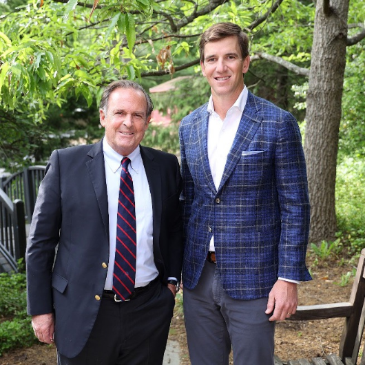 Peter Cancro and Eli Manning stand side-by-side and smile at the camera with greenery behind them.
