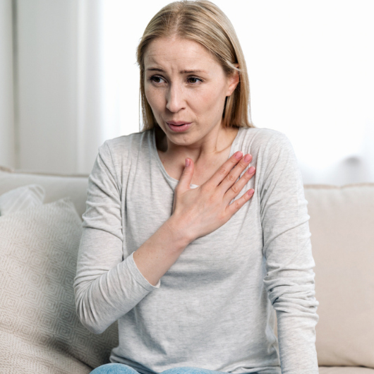 Young woman holds her hand on her chest and has difficulty breathing while sitting on a couch at home.