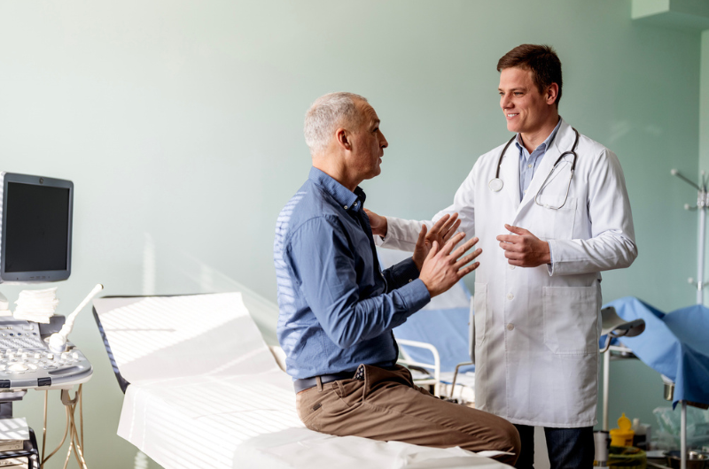 A male doctor stands and listens to his male patient talk.