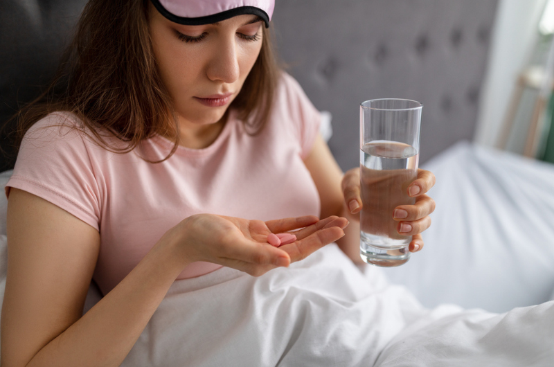 Caucasian woman sits up in her bed, holding a glass of water, looking at a melatonin pill.