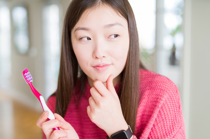 An Asian woman is deep in thought while holding a toothbrush.