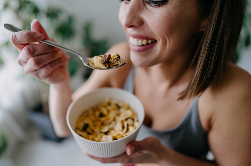 Cheerful young woman in sportswear smiling while enjoying a bowl of cereals with raisins.