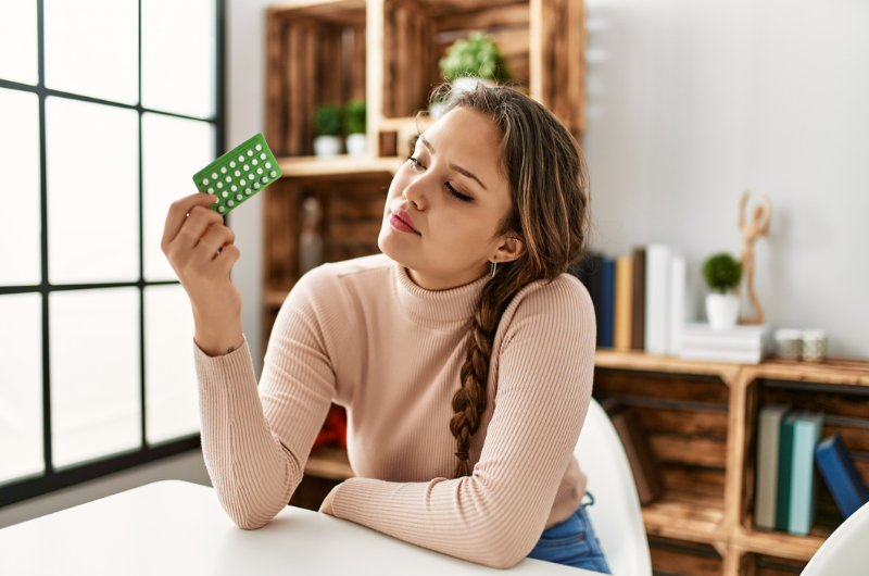Young, beautiful, Hispanic woman holding birth control pills sitting at a table at home.