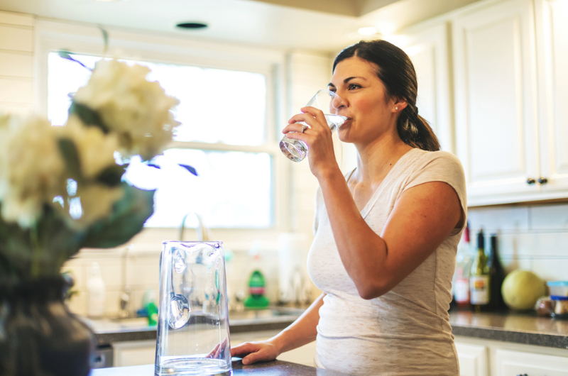 A young brunette woman drinks a glass of water in a kitchen to support healthy blood flow.