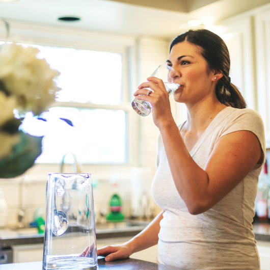 A young brunette woman drinks a glass of water in a kitchen to support healthy blood flow.
