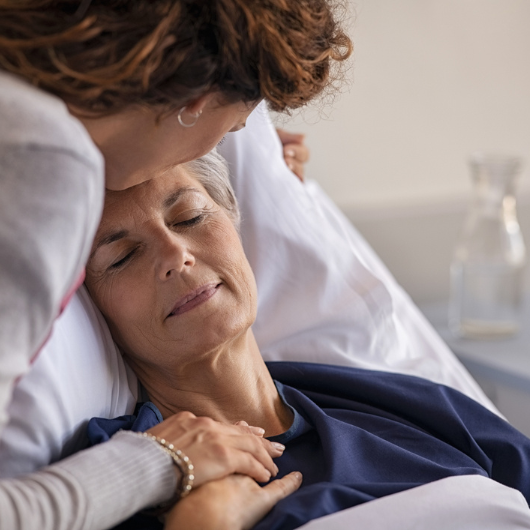 Close-up of adult daughter visiting and kissing her sick mother on the forehead while she rests in a hospital bed.