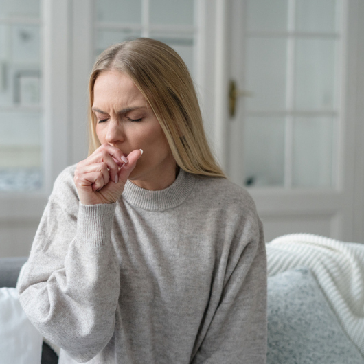 Blonde woman sits on a couch and coughs into her closed hand.