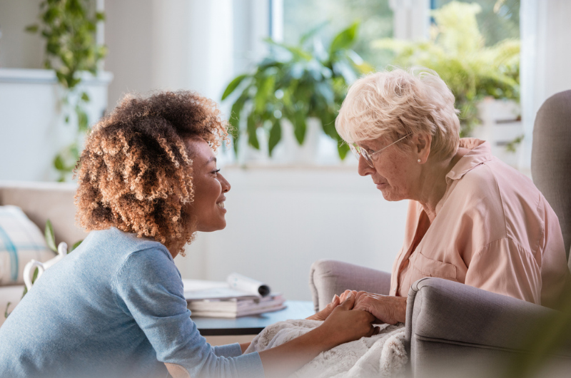 African American female home nurse talks with a senior woman, sitting in the living room and holding her hands.