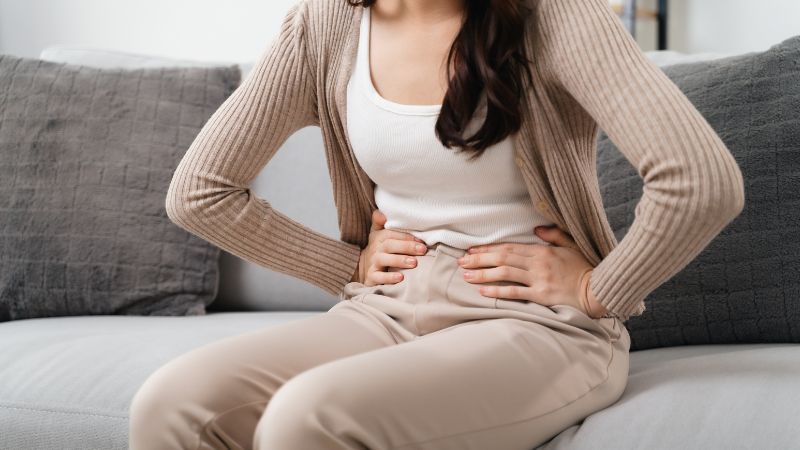 Image shows a close up of a woman holding her lower stomach while sitting on a couch.