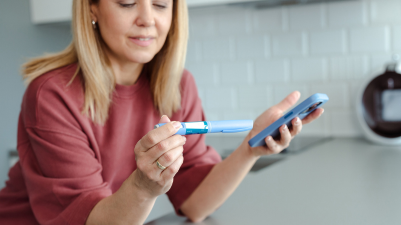 A woman holding a insulin pen and a smartphone, appearing focused and analytical, in a modern home kitchen interior.