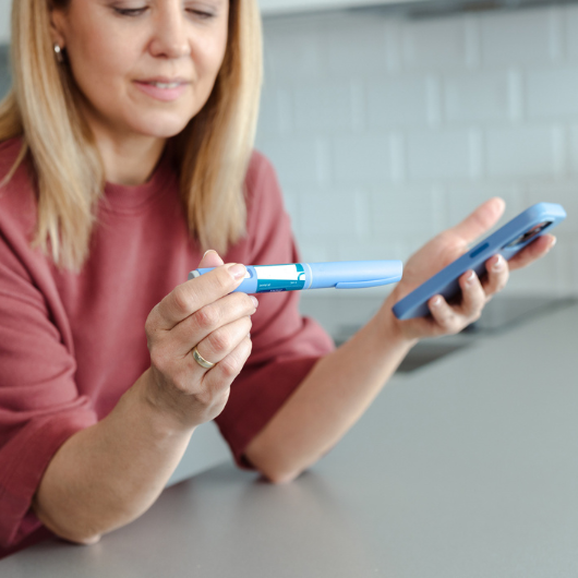 A woman holding a insulin pen and a smartphone, appearing focused and analytical, in a modern home kitchen interior.