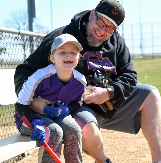 Russell and his daughter Devyn at the baseball field, sitting and smiling together.