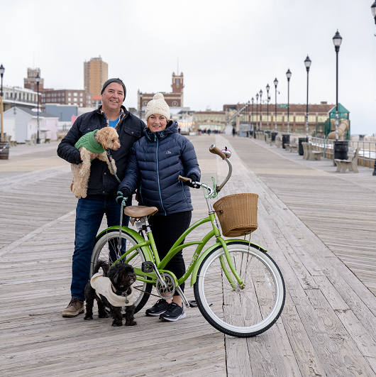 Cathy and her husband standing on the boardwalk in Asbury Park with her two dogs and bicycle.