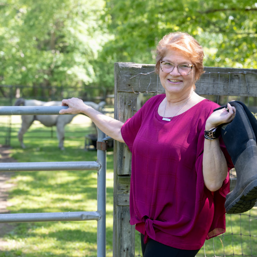 A woman holds onto a pair of boots while leaning up against the gate of a horse pen.