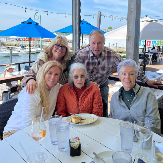 While dining outdoors, an elderly woman and her family smile at the camera.