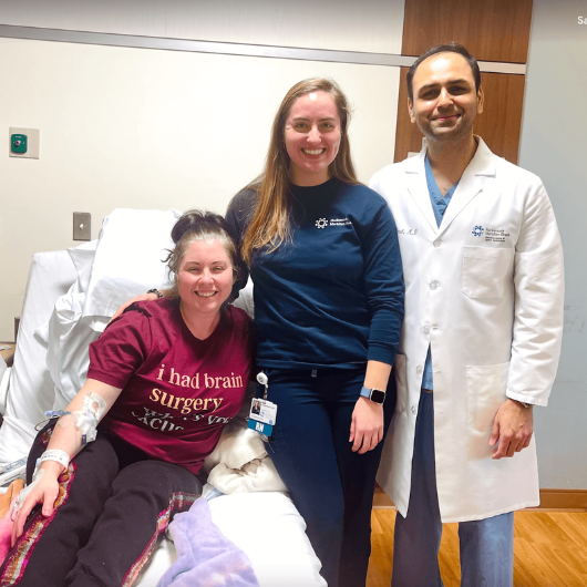 Selena Campione, 36, sits in a patient bed with brain surgeon, Nitesh Patel, M.D., standing beside her.
