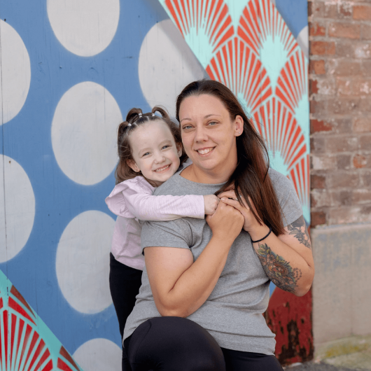 With her young daughter standing behind her, Lisa Ruding kneels and smiles.