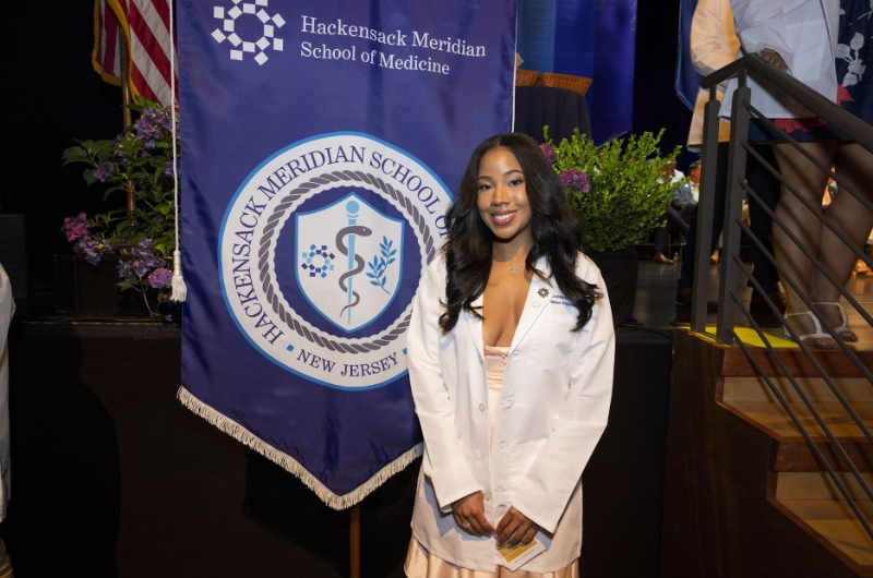 A young woman named Aya Taylor smiles next to a Hackensack Meridian School of Medicine banner.
