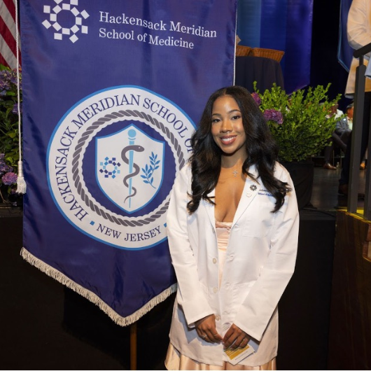 A young woman named Aya Taylor smiles next to a Hackensack Meridian School of Medicine banner.
