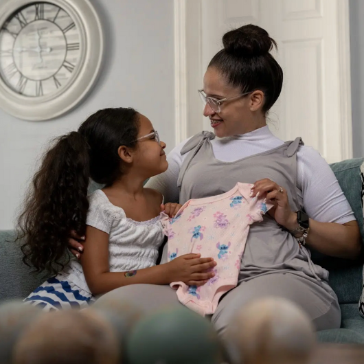 Swanny Fernandez holds a baby onesie while sitting on the couch next to a little girl.