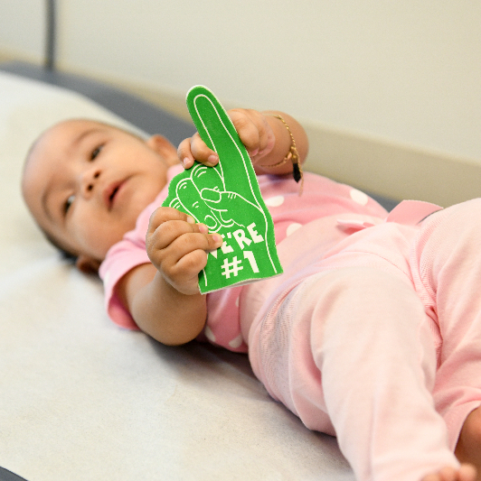 A baby laying, holding a green #1 foam finger.