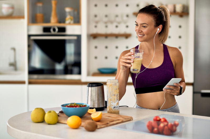 Happy, athletic woman drinks a smoothie while using a mobile phone and listening to music in the kitchen.